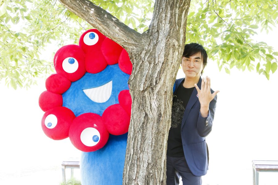 木を真ん中に右側から顔を出す河森プロデューサーと左側から顔を出すミャクミャク