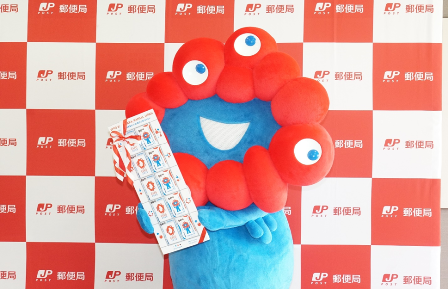 郵便局の赤と白の格子柄をバックに大きな特殊切手を持っているミャクミャク