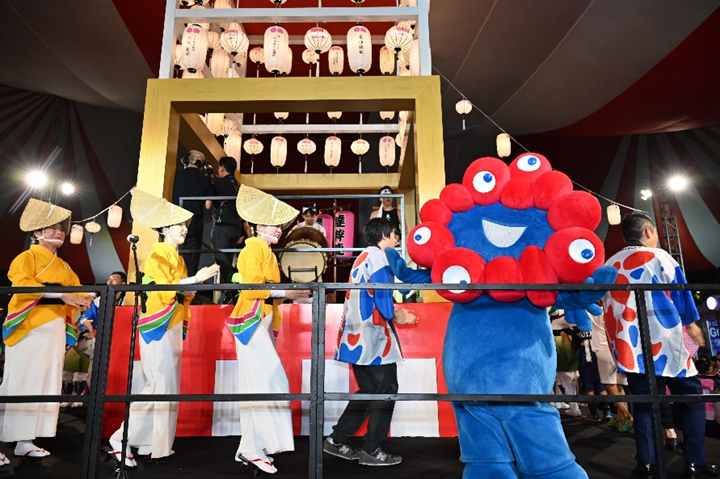 提灯が灯る櫓の前でミャクミャクと踊り子が楽しそうに踊っています。